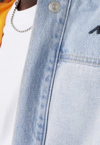 Jasnoniebieska kurtka dżinsowa z srebrnym guzikiem, pod którą znajduje się biały t-shirt oraz widoczny łańcuszek naszyjnik. Minimalistyczny design.