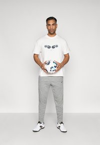 T-shirt branca com estampa gráfica, calças de fato cinzentas e sapatilhas atléticas brancas/prateadas. Segurando uma bola de futebol estampada. Fundo simples.