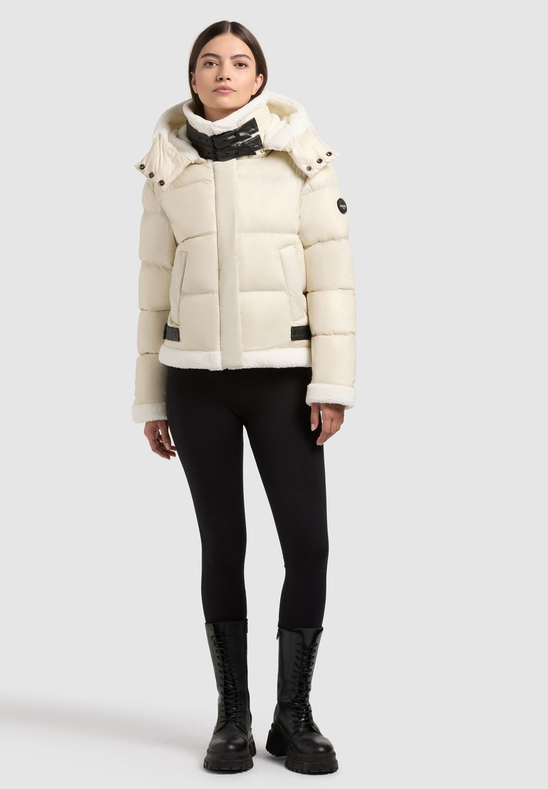 White puffer jacket with a textured, fur-lined hood, front pockets, and black accents. Model pairs it with black leggings and combat boots.