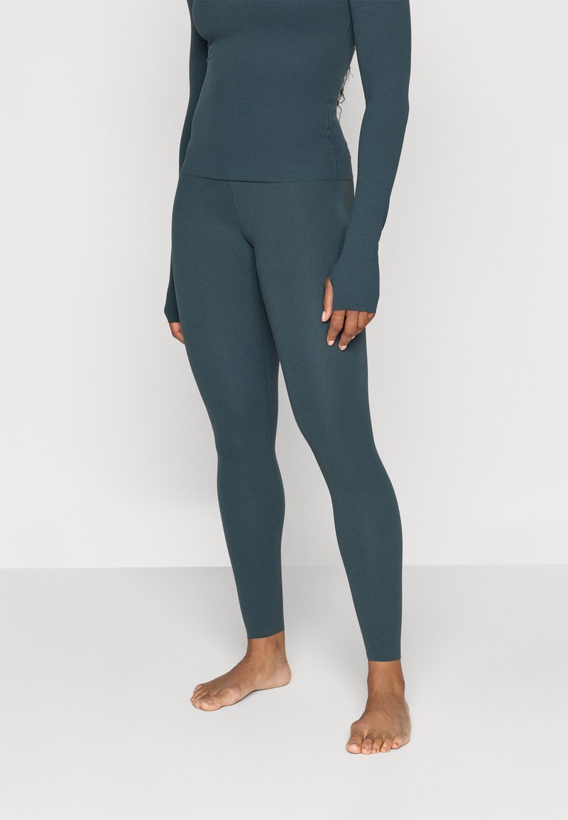 Mörk teal åtsittande långärmad topp och hjälpbyxor gjorda av stretchigt material. Tumhål och sömlös design. Barfotaställning, neutral bakgrund.