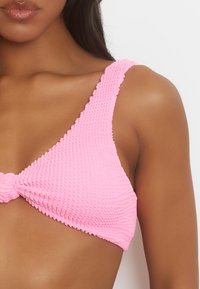 Top bikini rosa con tessuto testurizzato, dotato di spalline larghe e dettagli annodati frontalmente. Forma a triangolo con finitura liscia.