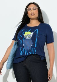 Laivastonsininen lyhythihainen t-paita, jossa on kuvio kissasta, jolla on keltainen hattu, sekä sininen ja valkoinen teksti. Pehmeä, kevyt kangas ja rento istuvuus.