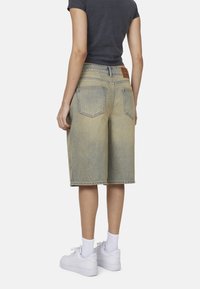 Shorts in denim chiaro con taglio dritto, dotati di un'etichetta sul retro, due tasche posteriori e orlo grezzo. Indossati con sneakers bianche.