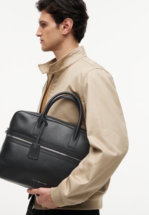 Hombre con chaqueta beige sosteniendo un maletín de cuero negro con cremallera plateada, de pie de lado sobre fondo blanco.