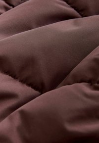 Pruun polsterdatud kangas, millel on sile, mati tekstuur, nähtavad õmblusread ja pehmed, quiltitud sektsioonid, mis loovad geomeetrilise mustri.