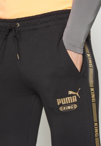Černé tepláky vyrobené z příjemného bavlněného směsového materiálu, s pozlaceným logem "PUMA KING", elastickým pasem na šňůrku a bočními kapsami s kontrastními pruhy.