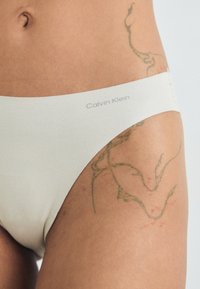 Lenjerie de femei Calvin Klein dintr-un material bej moale. Prezintă o talie netedă cu numele brandului. Include detalii vizibile de tatuaj verde în apropiere.