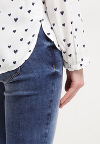 Blouse blanche à manches longues avec des motifs de cœurs noirs, rentrée dans un jean en denim bleu, vue latérale du vêtement et de la main droite.