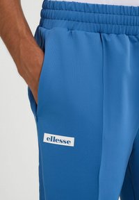 Niebieskie spodnie sportowe wykonane z gładkiego materiału, z elastycznym paskiem w talii, bocznymi kieszeniami i białą łatką z logo na udzie.