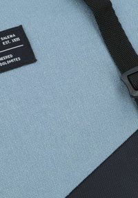 Blauem Stoff mit einer strukturierten Oberfläche, versehen mit einem schwarzen Etikett mit weißem Text und einem schwarzen Riemen, der teilweise an der Seite sichtbar ist.