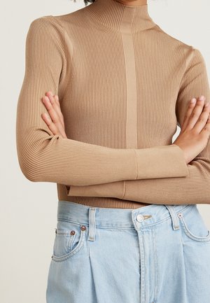 Personne portant un pull à col roulé côtelé beige à manches longues, bras croisés, associé à un jean taille haute bleu clair.