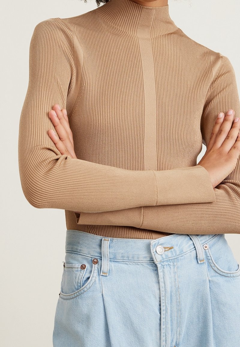 Personne portant un pull à col roulé côtelé beige à manches longues, bras croisés, associé à un jean taille haute bleu clair.