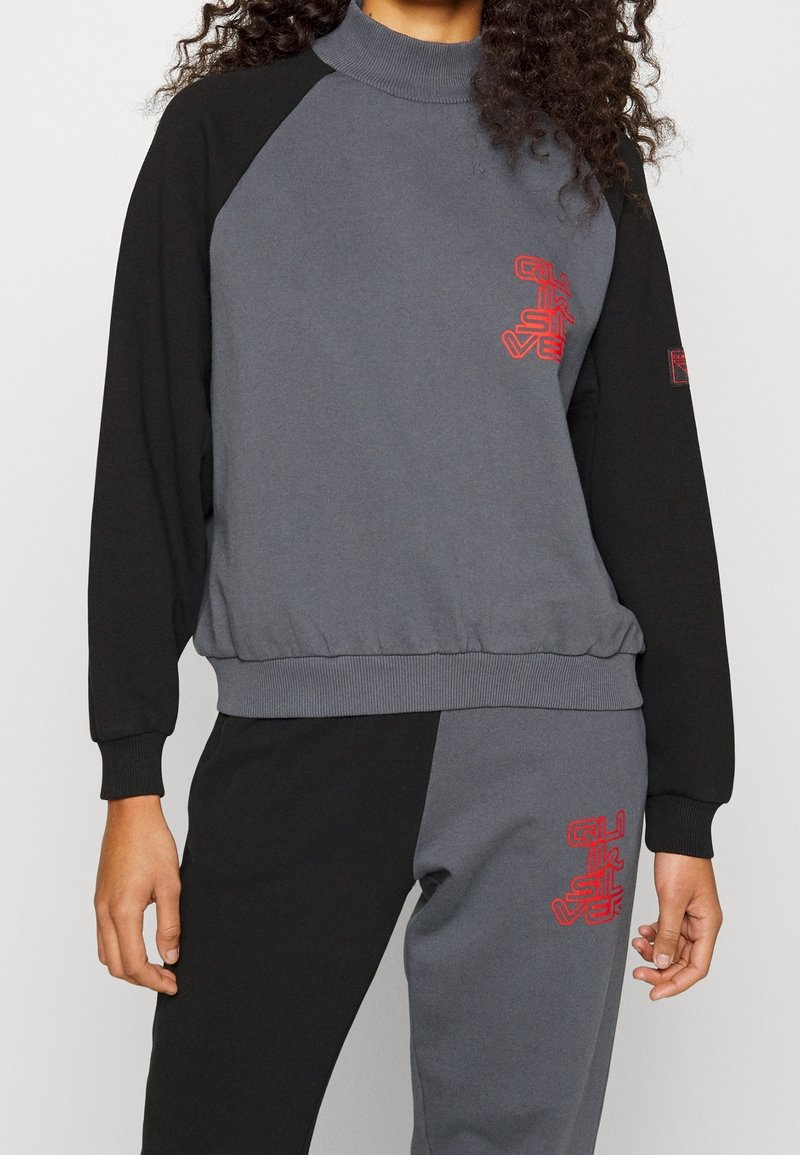 Grå och svart sweatshirt med hög krage, ribbad nederkant och mudd. Har röd logotypdetalj på bröstet och byxorna. Mjuk tygkänsla.