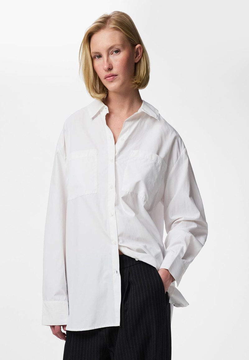 Femme blonde portant une chemise blanche boutonnée oversize et un pantalon noir à fines rayures, debout avec une main dans la poche, sur fond blanc.
