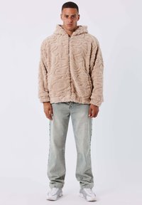 Giacca cappuccio beige e soffice con texture a zigzag, chiusura con zip e vestibilità ampia. Abbinata a jeans grigi chiari con bottoni a pressione sui lati.