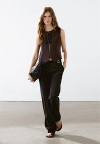 Zwarte cropped top met verticale plooien, gecombineerd met zwarte wijde broek. Model houdt een gestructureerde zwarte clutch vast en draagt platte sandalen.