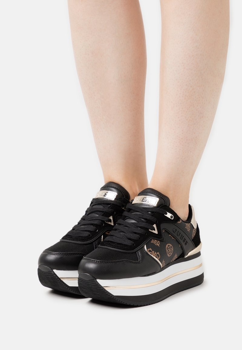 Jambes portant des baskets compensées noires Guess à lacets avec des motifs de logo marron et des semelles épaisses rayées blanc-doré sur fond blanc.