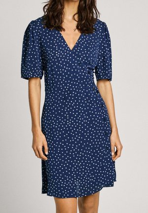 Granatowa sukienka w białe groszki, z kopertowym krojem, krótkimi bufkami oraz ozdobnym guzikiem wzdłuż boku. Gładka faktura tkaniny.