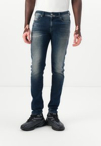 Sötétkék farmer skinny nadrág, elhalványult felülettel, elülső zsebekkel és enyhe kopásokkal. Fekete sportcipővel viselve.