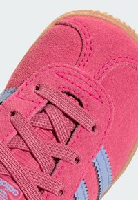 Sapatilha em camurça rosa com parte superior texturizada, atacadores planos e riscas azul claro com bordas em zigue-zague. Sola de borracha bege visível na parte inferior.