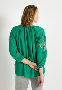 Blouse verte à manches longues avec un dos froncé, tissu texturé et détails en dentelle complexe sur les manches. Matériau doux et léger.