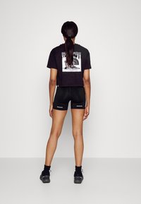 Černé croptop tričko s grafickým potiskem na zádech, spojené s černými přiléhavými šortkami s reflexními detaily. Obuv je tmavě šedá.
