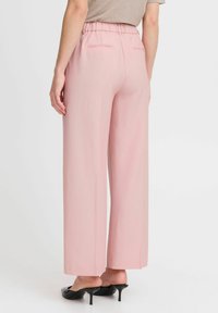 Pale pink wide-leg trousers with a gathered elastic waistband, two back pockets, and a smooth texture. Worn with black high-heeled mules.