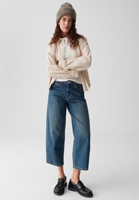 Donna con un berretto di maglia beige, camicia di corduroy crema sopra una maglietta bianca, jeans blu a vita alta e scarpe nere, in piedi con le braccia incrociate.
