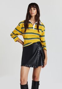 Frau in einem gelb- und grün gestreiften Langarmhemd, schwarzer Leder-Minikleid mit metallischen Ösen und schwarzen kniehohen Stiefeln, stehend.