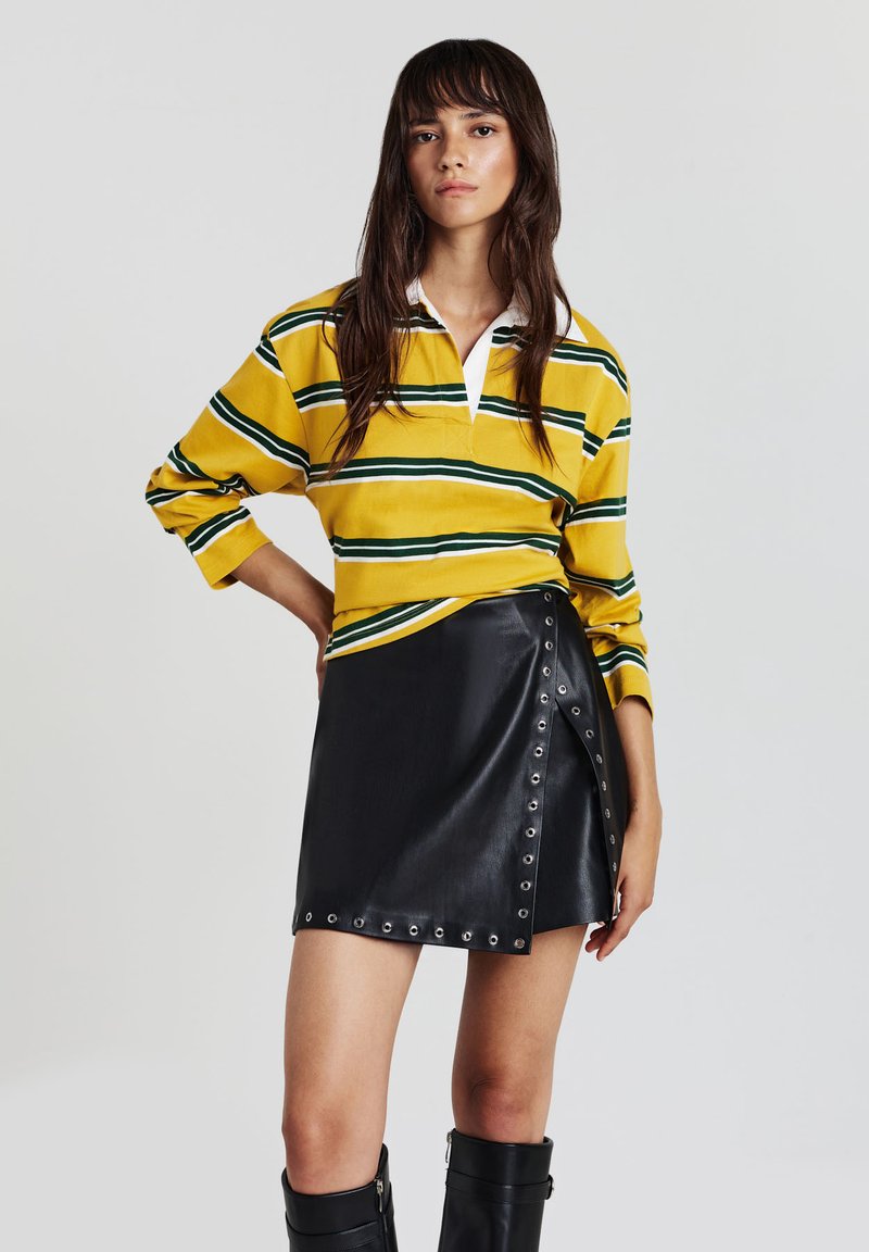 Frau in einem gelb- und grün gestreiften Langarmhemd, schwarzer Leder-Minikleid mit metallischen Ösen und schwarzen kniehohen Stiefeln, stehend.