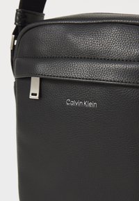 Borsa a spalla in pelle nera con finitura testurizzata, accessori in argento e logo "Calvin Klein" in bella vista.