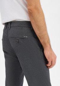 Graue strukturierte Hose mit schmaler Passform, Gesäßtaschen mit Knöpfen und einem kleinen metallischen Logo-Akzent nahe dem Bund.