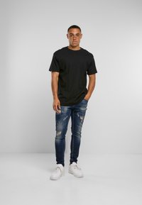 Mladý muž stojící čelem vzad, oblečený v černém tričku, roztrhaných modrých džínách a bílých teniskách na jednobarevném šedém pozadí.