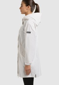 Donna con coda di cavallo scura che indossa una lunga giacca bianca con cappuccio e polsini elasticizzati, in piedi di lato su uno sfondo chiaro uniforme.