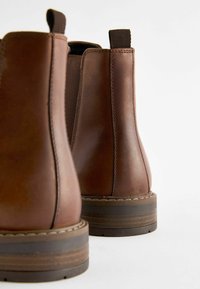 Botas de tobillo de cuero marrón con una textura suave, paneles laterales elásticos y suelas de goma marrón con un diseño de rayas horizontales.