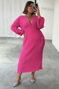 Long pink dress with a V-neck, long sleeves, and subtle gold heart patterns. Soft pleated texture, knee-length with a scalloped hem. Silver heels.