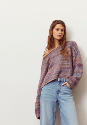 Maglione oversize multicolore con lavorazione a maglia testurizzata, indossato sceso su una spalla, abbinato a jeans azzurri a vita alta. Sfondo semplice.