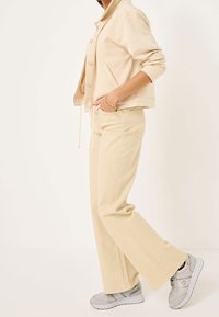 Veste beige à boutons, associée à un pantalon large beige clair et à des baskets grises. Tissu lisse avec une coupe décontractée et des poches.