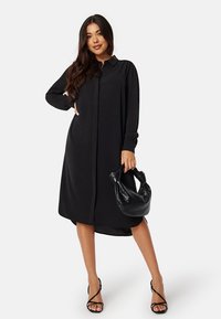 Abito camicia nero con maniche lunghe, chiusura a bottoni e silhouette rilassata. Abbinato a una borsa nera testurizzata e tacchi con cinturini.