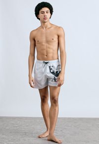 Ανδρικό γκρι μαγιό βερμούδα με μαύρο γραφικό σχέδιο, ελαστική μέση και κορδόνι. Ελαφρύ ύφασμα, μήκος μέχρι το μέσο μηρού, λεία υφή.