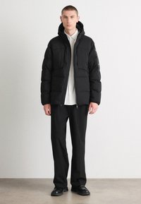 Jachetă puffer neagră cu glugă, având un design matlasat, buzunare frontale și închidere cu fermoar, asociată cu pantaloni negri largi și încălțăminte.