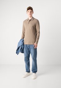 Camisa polo de manga longa castanha, colarinho caqui claro, jeans de ganga, tênis brancos, segurando uma jaqueta de ganga azul, fundo neutro.