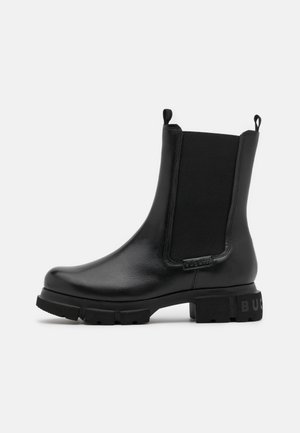 Bottine noire en cuir avec panneaux latéraux élastiques, languettes, semelle épaisse et logo discret embossé au talon et sur le côté.