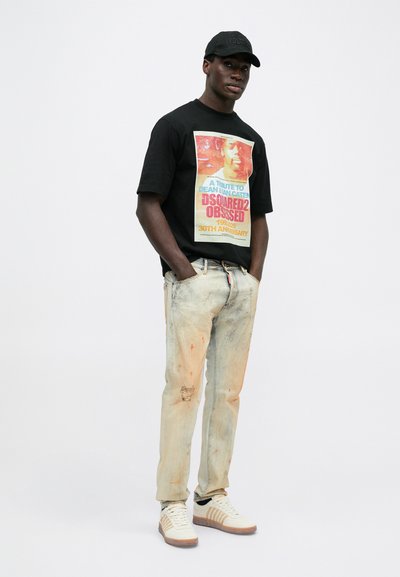 Tricou negru cu un print graficColorat. Blugi ușor uzați, cu pete portocalii și o ruptură la genunchi. Adidași albi cu tălpi maro.