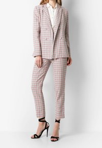 Kvinde iført en rød og hvidternet blazer med matchende bukser, hvid skjorte med knapper og sorte højhælede sandaler, stående mod en hvid baggrund.