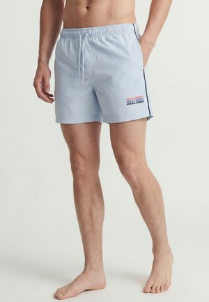 Bañador corto de hombre azul claro con cintura elástica, cordón, bolsillos laterales y logo "Jack & Jones" en la pierna izquierda, usado descalzo.