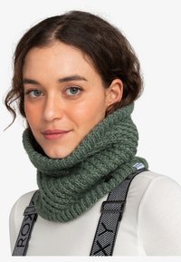Strikket grøn cowl-halsvarmer lavet af tykt garn med en struktureret ribmønster, der har en knapdetalje og et mærke på siden.