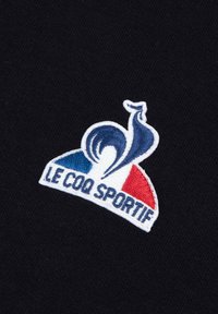 Logo ricamato di Le Coq Sportif su uno sfondo nero; presenta accenti blu e rossi con un design a spirale.