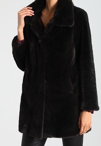 Kvinna iklädd en svart fuskpälsjacka med bred krage, långa ärmar och sidofickor över en mörk klädsel.