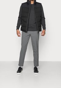 Veste rembourrée noire portée sur un pull gris, associée à un pantalon gris clair et des baskets noires. Textures lisses, coupe standard, sans motifs visibles.